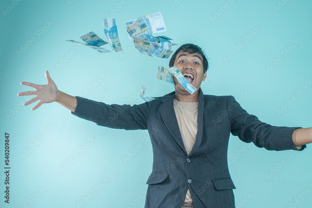 A successful Asian businessman wearing a black suit is throwing money away so the money is flying on a blue background A successful Asian businessman wearing a black suit is throwing money away so the money is flying on a blue background