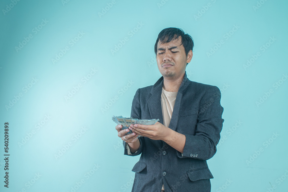 Successful Asian businessman wearing a black suit looking at his meager money with a sad expression standing on blue background Successful Asian businessman wearing a black suit looking at his meager money with a sad expression standing on blue background