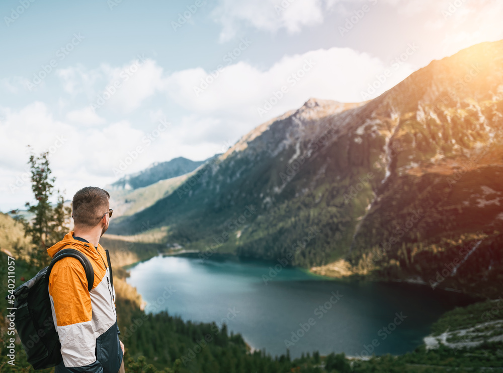 Man traveler smiles with beatufil mountain lake and forest in the background. Concept of outdoor adventures and hiking. Bearded young caucasian man wears backpack and sport outfit in the mountains. Man traveler smiles with beatufil mountain lake and forest in the background. Concept of outdoor adventures and hiking. Bearded young caucasian man wears backpack and sport outfit in the mountains.