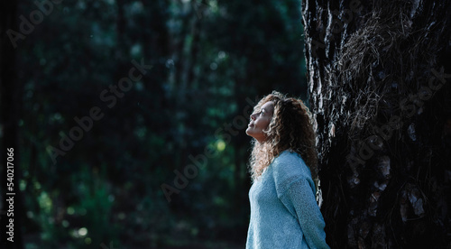 Dark image of woman standing against a tree trunk in oudoor nature leisure activity with woods in background. One female people enjoy forest looking up and relaxing. Environment in the park. photo