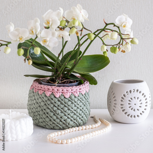 Pearl necklace and beads in pink crocheted basket, white candle, candleholder and lisianthus bouquet on white table in white interior. Cozy home atmosphere, Handicraft advertisement photo