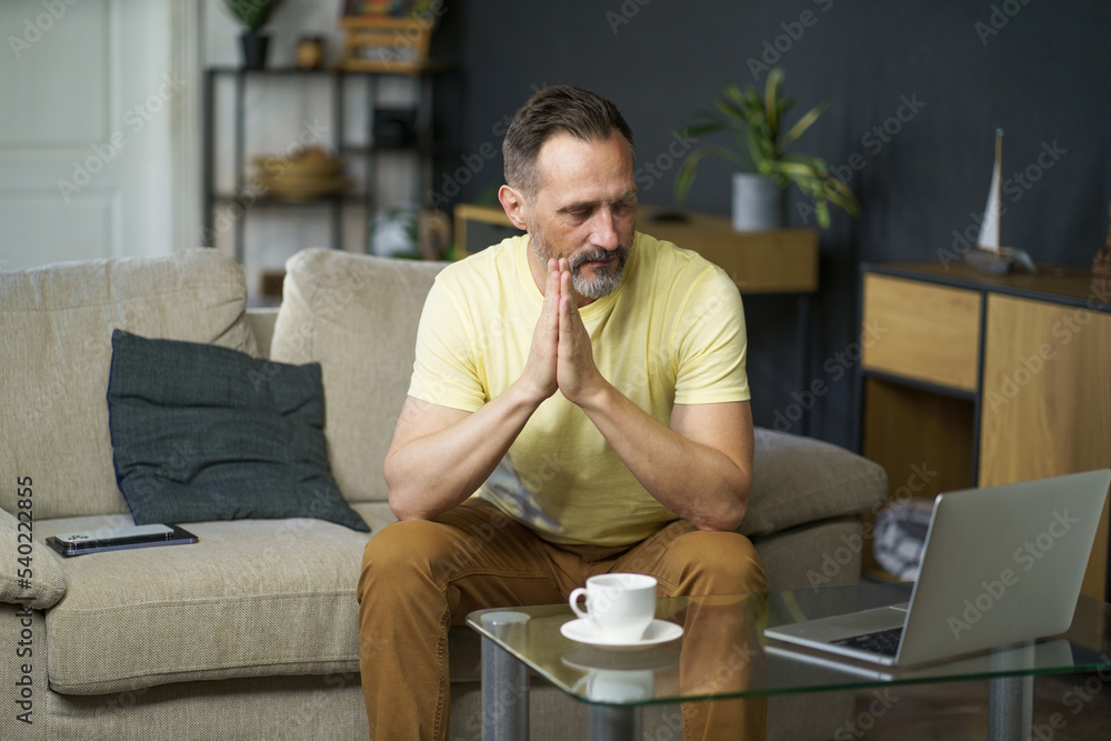 Emotional stress mature man sits on sofa with hands put together next to coffee table with laptop and cup on it in home interior wearing casual. Middle age crisis, devours, lonely, puzzled man Emotional stress mature man sits on sofa with hands put together next to coffee table with laptop and cup on it in home interior wearing casual. Middle age crisis, devours, lonely, puzzled man