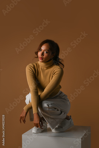 Trendy young woman wearing warm sweater sitting on orange background. Fashion studio photo, Autumn concept
