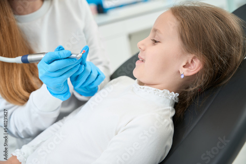 Pediatric dentist telling about teeth-polishing procedure to smiling little girl