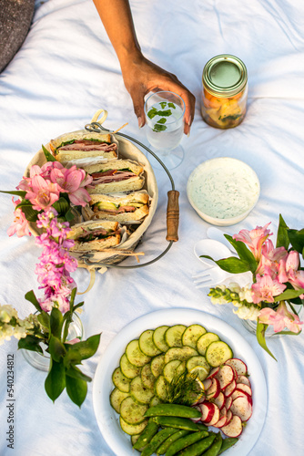 Black woman holding a glass of mint water while enjoying a luxury picnic in the park with fresh flowers, sandwiches, crudites platter and fruit salad