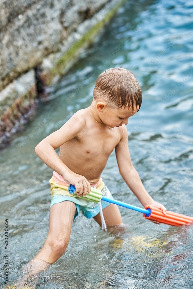 Small happy child plays with water-jet gun standing in clear blue shallow water Small happy child plays with water-jet gun standing in clear blue shallow water