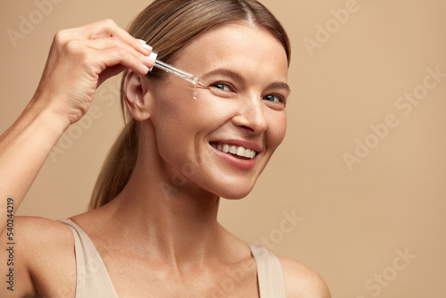 Woman Dropping Serum on Face. European Girl Applying Serum Collagen Moisturizer from Pipette on Face close up. Positive Laughing Beautiful Woman. Concept of Face Skin Care  photo