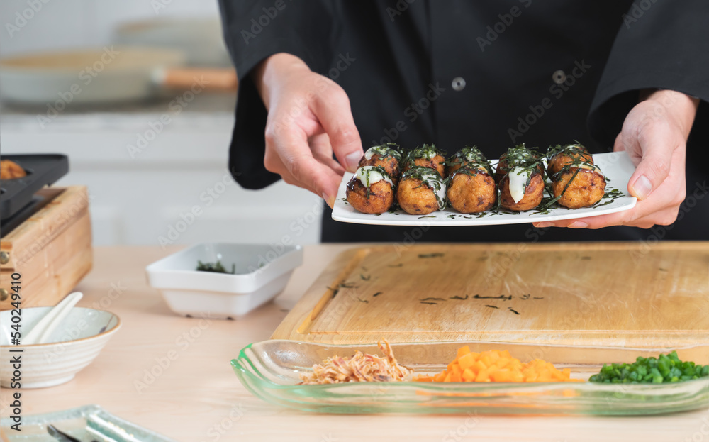 Selective focus on japanese food called takoyaki in plate from hot pan in hands of anonymous young chef man, in black uniform, showing dishes on table at kitchen restaurant