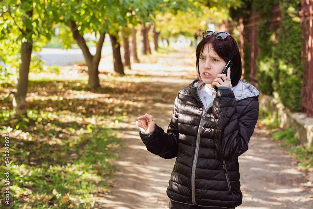 A teenage girl in an autumn park is angrily talking to someone on the phone and waving hands, arguing with dad on the phone, not wanting to listen to her dad's instructions A teenage girl in an autumn park is angrily talking to someone on the phone and waving hands, arguing with dad on the phone, not wanting to listen to her dad's instructions