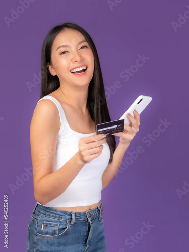 Beautiful Asian brunette woman cute girl in white tank top holding credit card and smartphone ,mobile phone uses a online banking for pay online shopping isolated on purple background