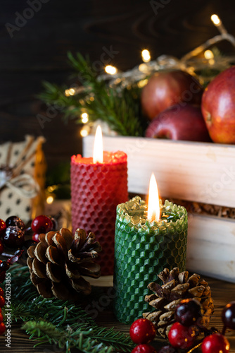 Red and green candle made of natural honeycomb on a wooden table. Red berries, cones and spruce branches in the background. Dark wooden background