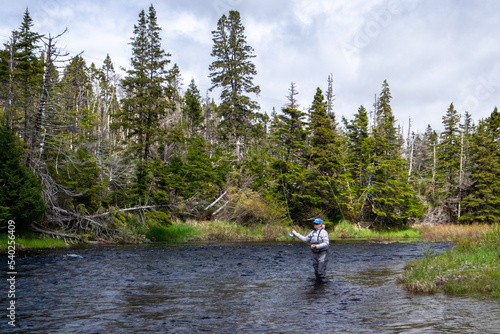 A middle age female stands in a large salmon river casting a fishing rod holding line in her left hand. The blonde long haired lady is wearing a blue hat, white t-shirt, and waders fly-fishing. 