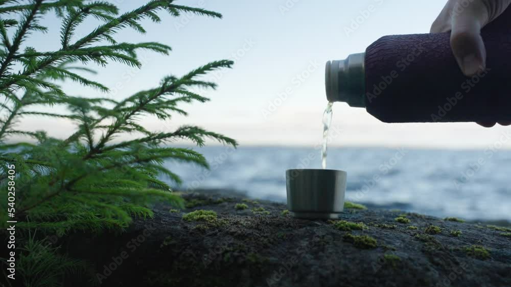 Thermos pours hot tea into an aluminum cup close-up against the background of northern nature in autumn Thermos pours hot tea into an aluminum cup close-up against the background of northern nature in autumn