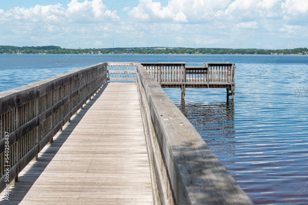 A long wooden boat wharf. The grey worn plank style pier juts out into a lake on all three sides with tree covered land in the background under blue sky with clouds. The water is calm and blue.  
