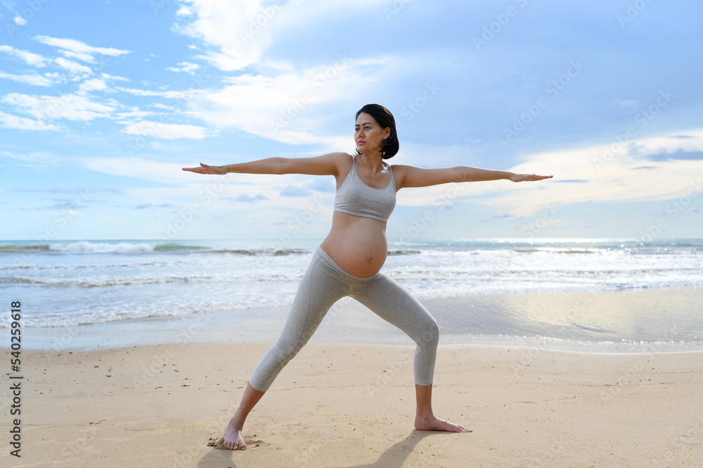 Pregnant woman practices yoga and meditation while doing warrior two pose stand on peaceful beach with nature scene- Healthy lifestyle concept Pregnant woman practices yoga and meditation while doing warrior two pose stand on peaceful beach with nature scene- Healthy lifestyle concept