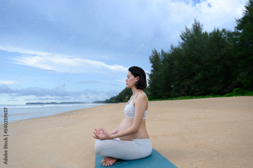 Pregnant woman practices yoga and meditation while doing lotus poses and sits on the peaceful beach with a nature scene- Healthy lifestyle concept