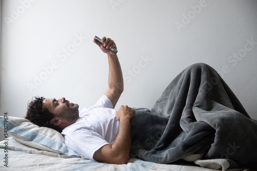 man lying on his bed holding his phone with both hands up, man working at his home lying on the bed