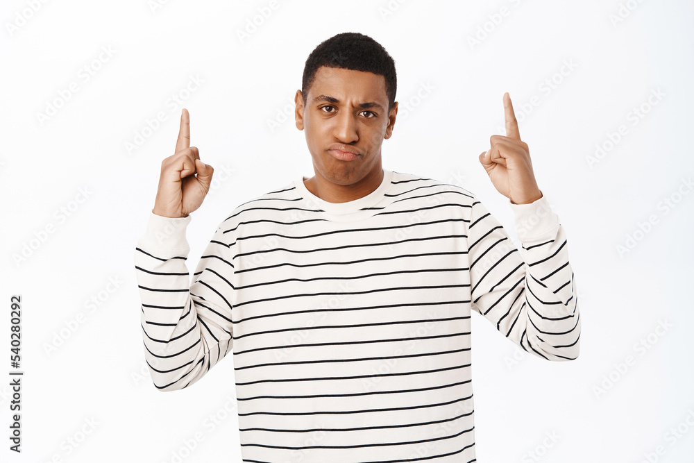 Annoyed african american man points up, sulks and looks bothered by smth, stands over white background Annoyed african american man points up, sulks and looks bothered by smth, stands over white background