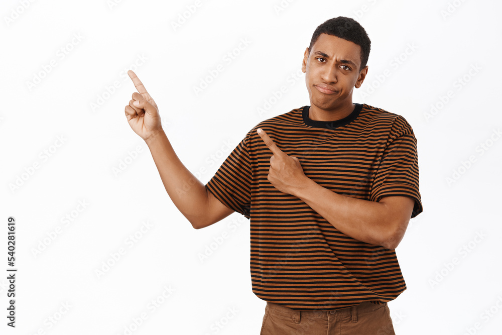 Portrait of disappointed african american man pointing finger left, frowning and looking upset, complaining at promotion text, white background Portrait of disappointed african american man pointing finger left, frowning and looking upset, complaining at promotion text, white background