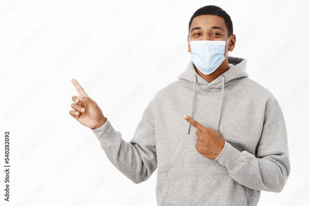 Covid awareness, healthcare concept. Portrait of african american guy in medical mask, pointing finger left, showing promotion text, advertisement, white background Covid awareness, healthcare concept. Portrait of african american guy in medical mask, pointing finger left, showing promotion text, advertisement, white background
