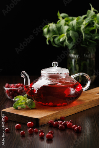 Red herbal tea with cranberries and mint in a glass teapot on a black background. A bunch of mint in the background. Cranberries in the foreground. Selective focus.