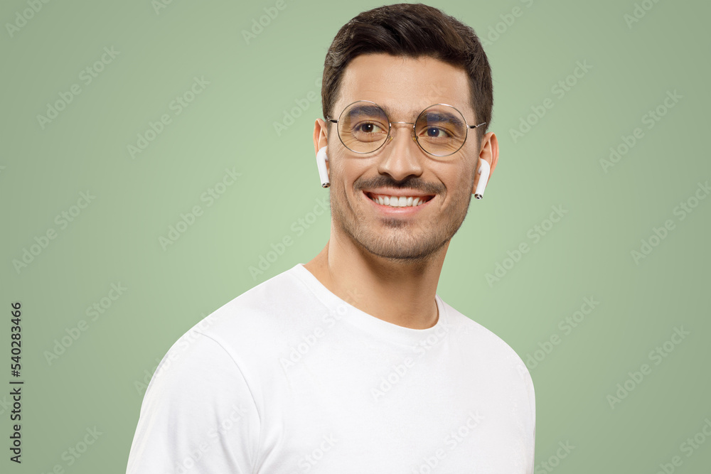 Handsome young man in white t-shirt, eyeglasses and wireless earphones, looking aside with smile Handsome young man in white t-shirt, eyeglasses and wireless earphones, looking aside with smile