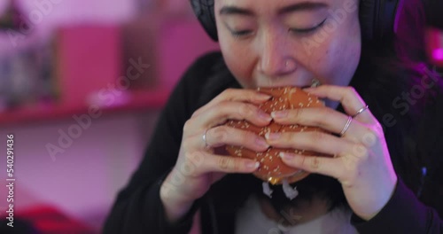 Gaming, fast food and asian woman eating burger and chips while playing live online cyber game on computer in Korea. Gamer girl, headphones and streaming esports subscription in chair and neon room photo