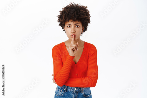 Beautiful young woman, 25 years, shushing, makes shh hush sign, stands with finger pressed to lips, taboo gesture, stands over white background