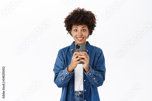 Portrait of modern girl takes photos on mobile phone, records video with her smartphone, stands happy over white background