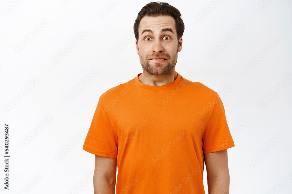 Fototapeta premium Image of guy looks with tempted, excited face, raising eyebrows up, standing in orange tshirt, white background