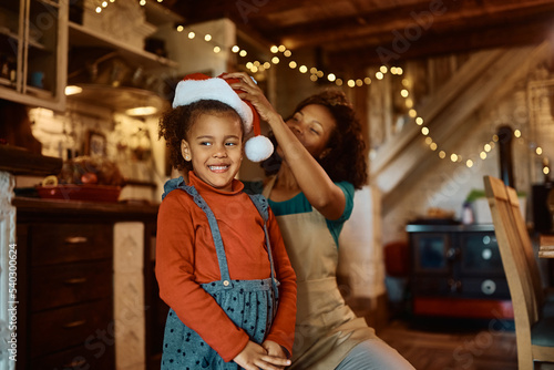 Happy black girl getting Santa hat from her mother while preparing food together in kitchen.