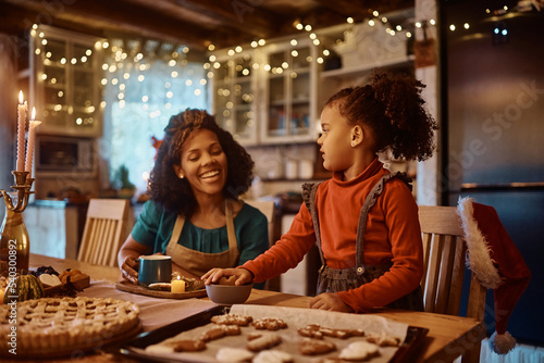 Black little girl assisting her mother in making gingerbread cookies for holidays in kitchen.