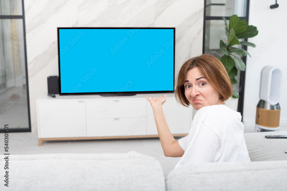 Shocked girl sitting on the couch, watching TV and pointing on screen. She demonstrating her disgusting. Empty blue screen and free space on television concept Shocked girl sitting on the couch, watching TV and pointing on screen. She demonstrating her disgusting. Empty blue screen and free space on television concept