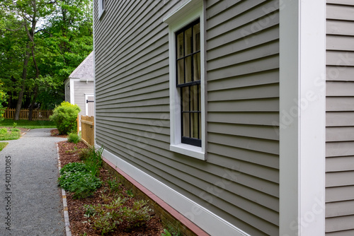 Old tan colored house with white trim and black wood window frame spacers. The glass in the window is wavy and reflects the sun. The exterior wall is made of vintage beige cape cod clapboard siding.