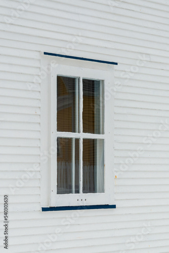 A small single casement window in a white narrow wood beveled clapboard siding exterior wall of a vintage house. The glass window has four panes with green narrow decorative trim on the outside edge. 