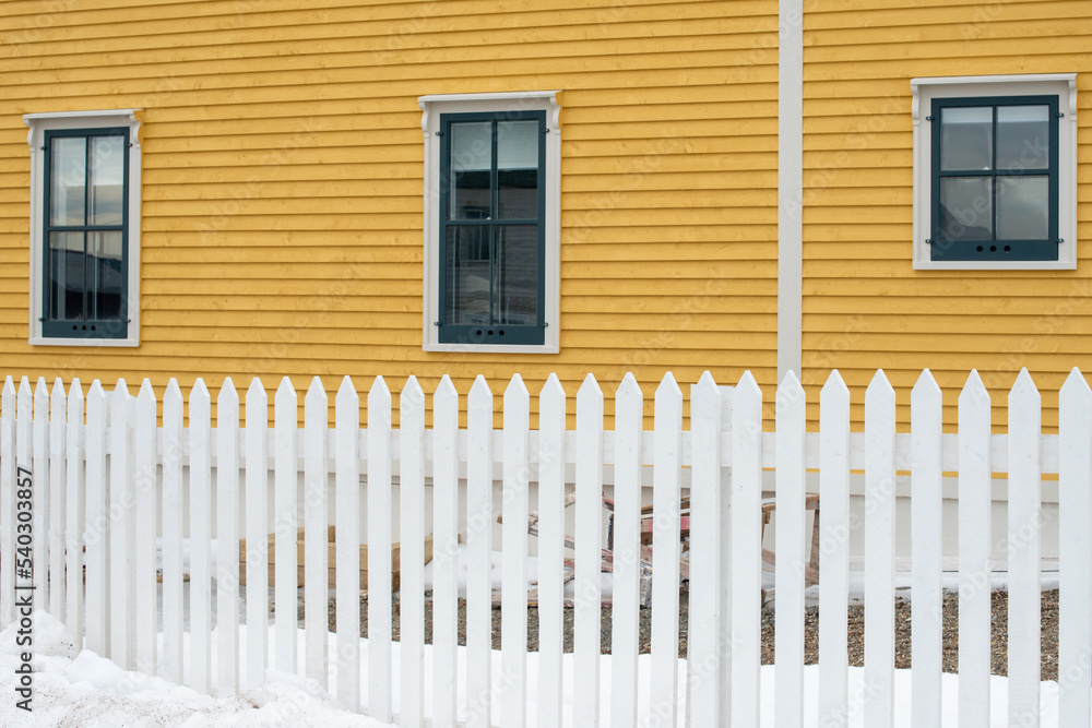 A white wooden picket fence is in the foreground with a bright yellow cape cod siding covered building in the background. The house has multiple vintage windows on the exterior wall with green trim. A white wooden picket fence is in the foreground with a bright yellow cape cod siding covered building in the background. The house has multiple vintage windows on the exterior wall with green trim.