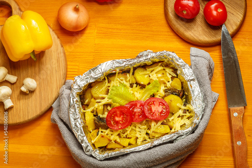 A cheese-baked card decorated with tomatoes on foil in the form on a wooden table next to vegetables and a knife.