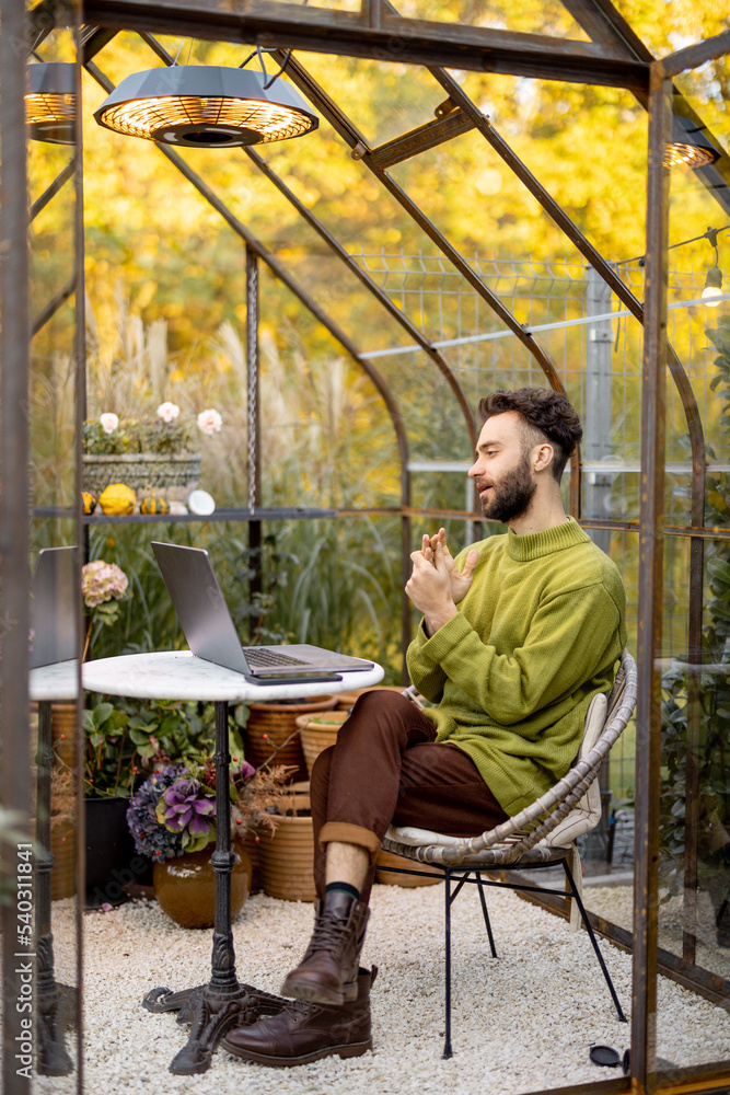 Man works on laptop, talking online while sitting by the round table in glasshouse with plants and flowers at backyard. Work from home at cozy atmosphere on nature