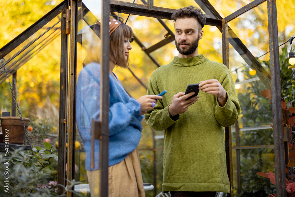 Young stylish man and woman talk together while standing with phones in front of tiny glass orangery in garden. Two hipsters hang out outdoors