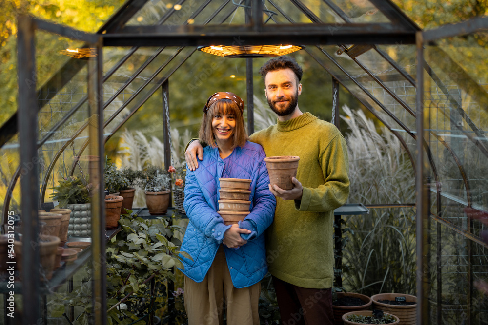 Obraz premium Portrait of a young couple stand together with clay jugs in small greenhouse for growing flowers in garden. Hobby or small family business of growing flowers concept