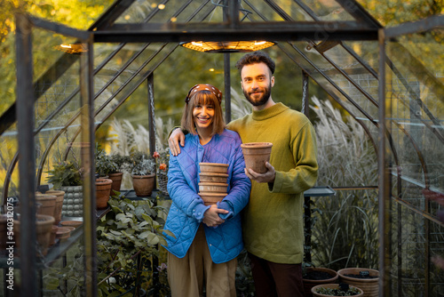 Portrait of a young couple stand together with clay jugs in small greenhouse for growing flowers in garden. Hobby or small family business of growing flowers concept