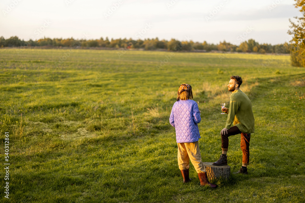 Young stylish couple hang out together, talking and drinking wine on green field during sunset. Spending autumn time outdoors