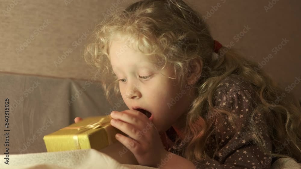 portrait of cute funny little girl in a festive cap opens a gift lying on the bed, christmas surprise, eve of christmas portrait of cute funny little girl in a festive cap opens a gift lying on the bed, christmas surprise, eve of christmas