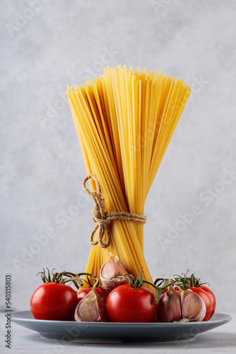 Spaghetti pasta with tomatoes and garlic on gray background. Dry italian pasta and vegetables on gray plate. Close-up