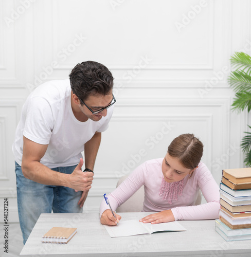 Father and daughter studying together online at home. He explaining lesson and she listening him carefully. Stack of books, laptop and exercise book on the desk. Online education concept