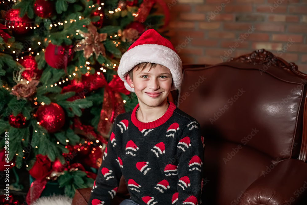 A funny boy in a Santa Claus hat on a vintage chair in a Christmas-decorated room. A funny boy in a Santa Claus hat on a vintage chair in a Christmas-decorated room.