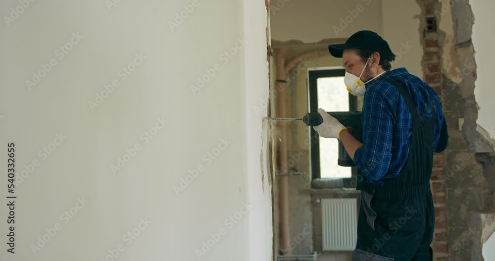 A man drills a hole in the wall of an apartment under renovation with a large drill. The construction worker, dressed in a protective suit, works with gloves. A man drills a hole in the wall of an apartment under renovation with a large drill. The construction worker, dressed in a protective suit, works with gloves.