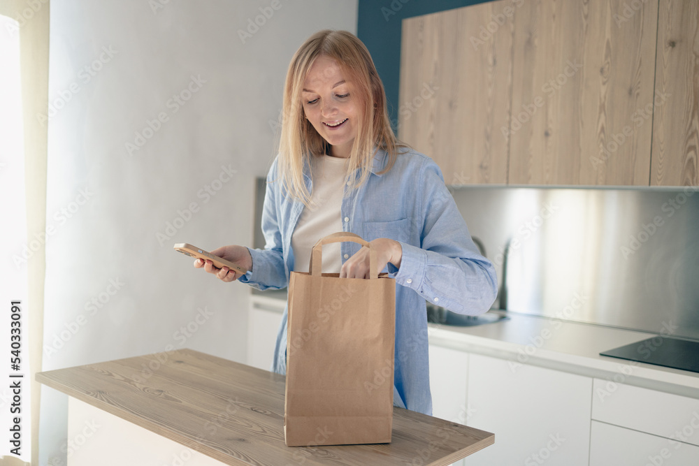 Food delivery concept. Woman packing parcel for sending, using smartphone, ordering delivery service