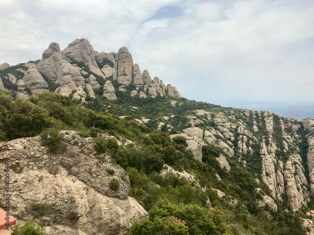 Montserrat, Spain, June 2019 - A rocky mountain with trees in the background with Montserrat in the background