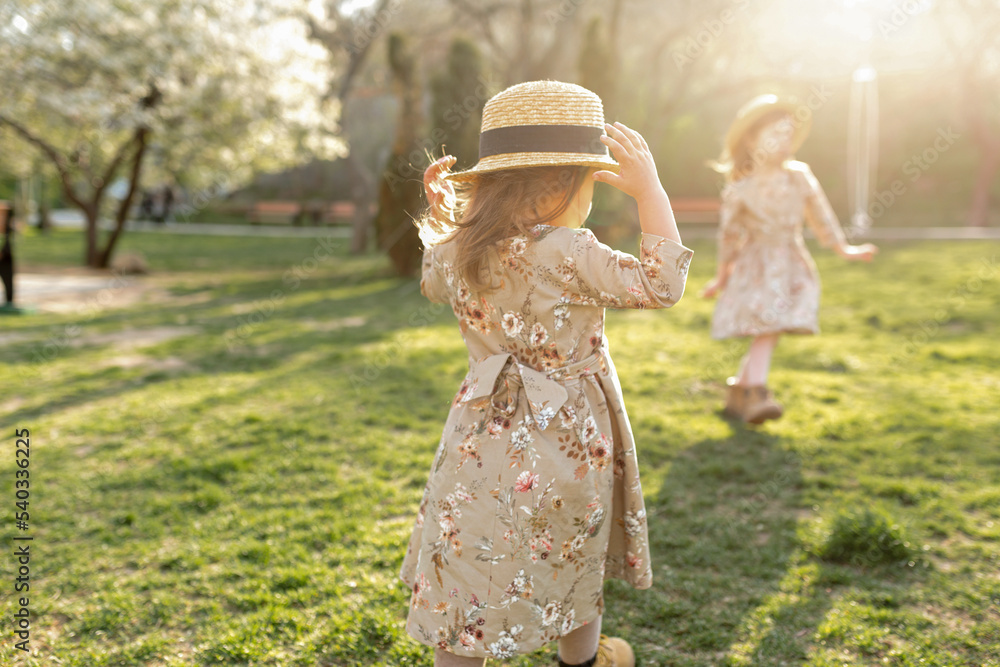 Outdoor portrait of cute little girl wearing spring dress in floral print and straw hat with ribbon having fun and running with her friend inn sunny warm autumn day